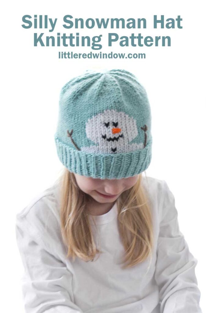 Blond girl in a white shirt wearing a light blue knit hat with a smiling snowman face peeking out over the folded brim smiling and looking down in front of a white background