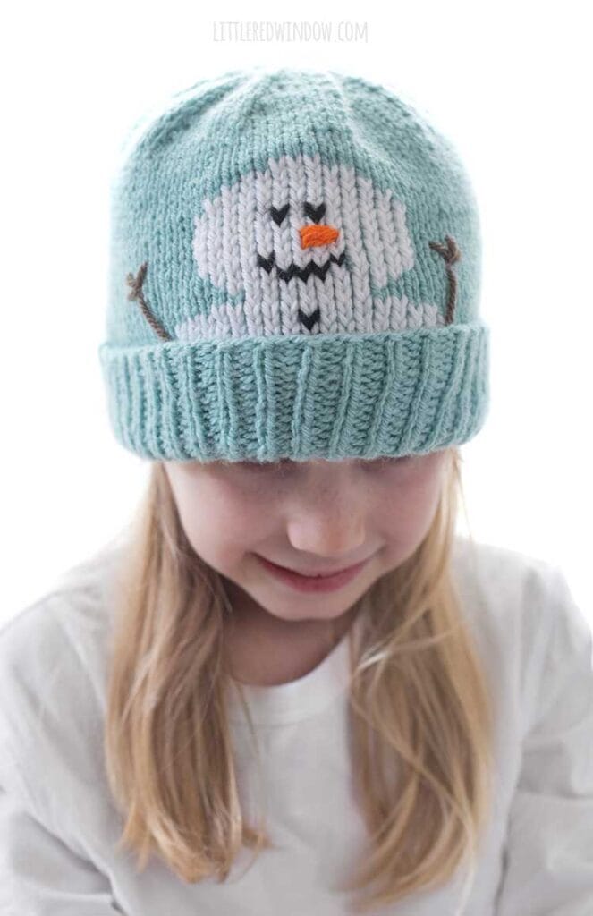 closeup of Blond girl in a white shirt wearing a light blue knit hat with a smiling snowman face peeking out over the folded brim smiling and looking down in front of a white background