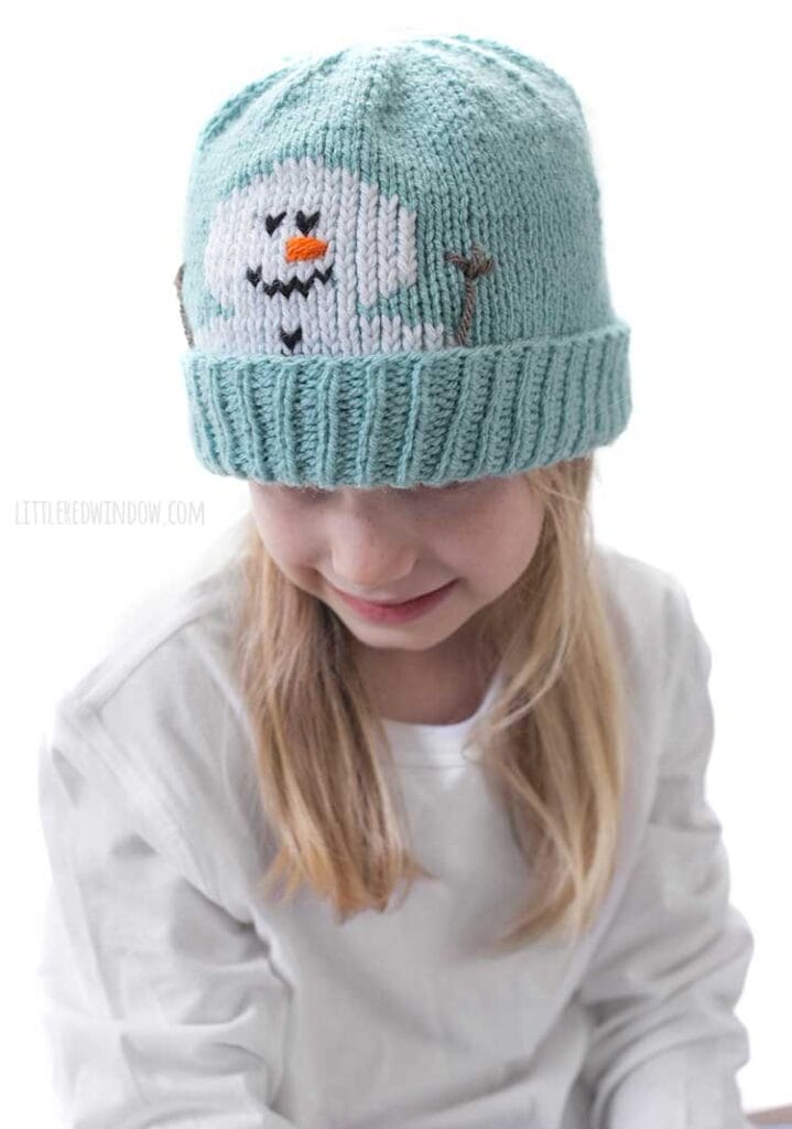 Blond girl in a white shirt wearing a light blue knit hat with a smiling snowman face peeking out over the folded brim smiling and looking down and to the left in front of a white background