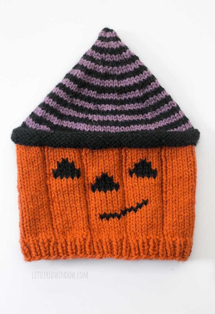 Flat lay of a knit hat that looks like an orange Jack o lantern with a smiling face wearing a striped witch's hat in front of a white background