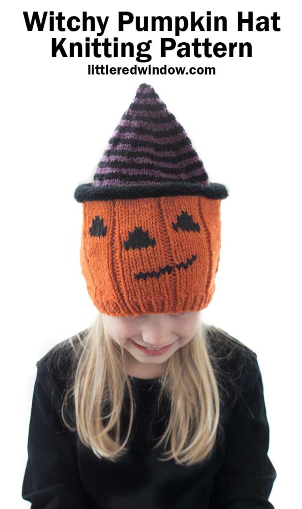 Smiling blond girl wearing a knit hat that looks like a jack o lantern wearing a purple and black striped witch hat while she is is looking down