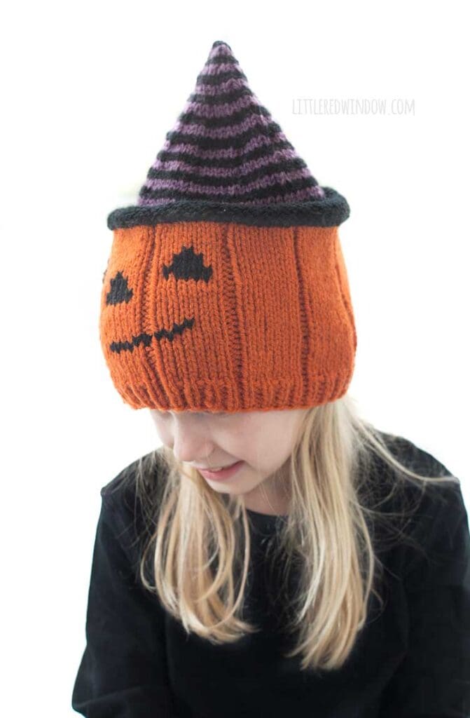 Smiling blond girl wearing a knit hat that looks like a jack o lantern wearing a purple and black striped witch hat while she is is looking down and to the left