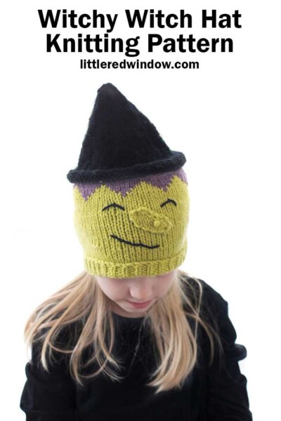 Little blond girl in a black shirt wearing a hat that looks like a witch including green witchy skin warty nose purple hair and black pointy witch hat while the girl is looking down and smiling