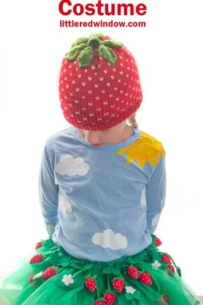 girl wearing a strawberry hat and green tutu skirt with felt strawberries all over it looking down at her lap