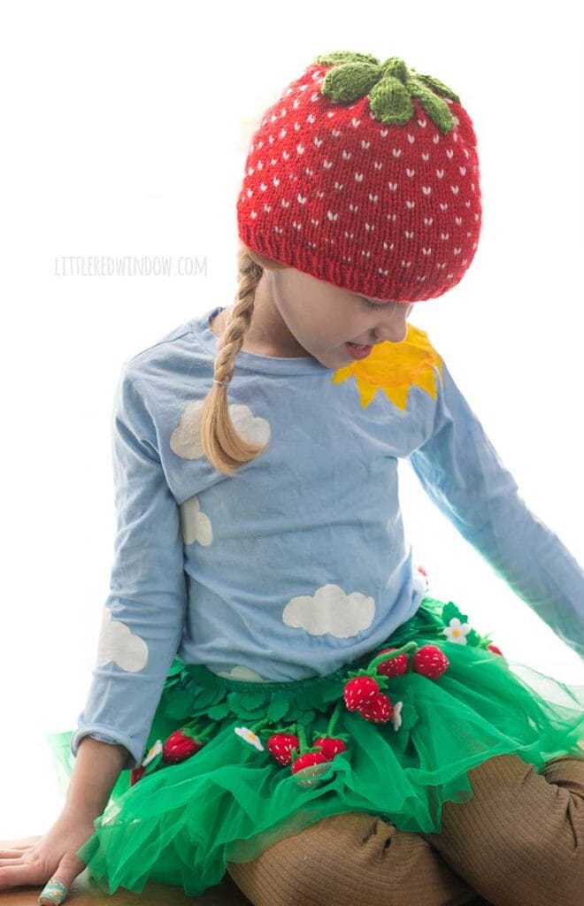 girl wearing a strawberry hat and green tutu skirt with felt strawberries all over it looking down and to the right