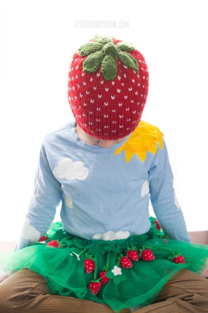 girl wearing a strawberry hat and green tutu skirt with felt strawberries all over it looking down at her lap