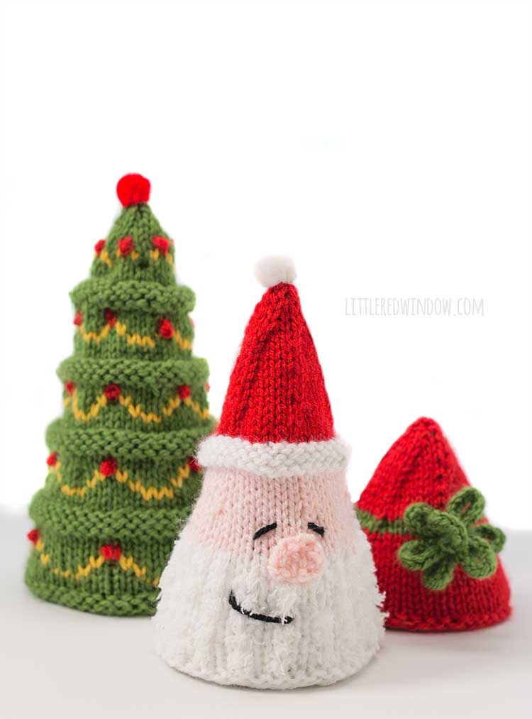 Three cone shaped knit Christmas trees that look like a classic green tree, a Santa head and a Christmas present with a bow in front of a white background the Santa is in front