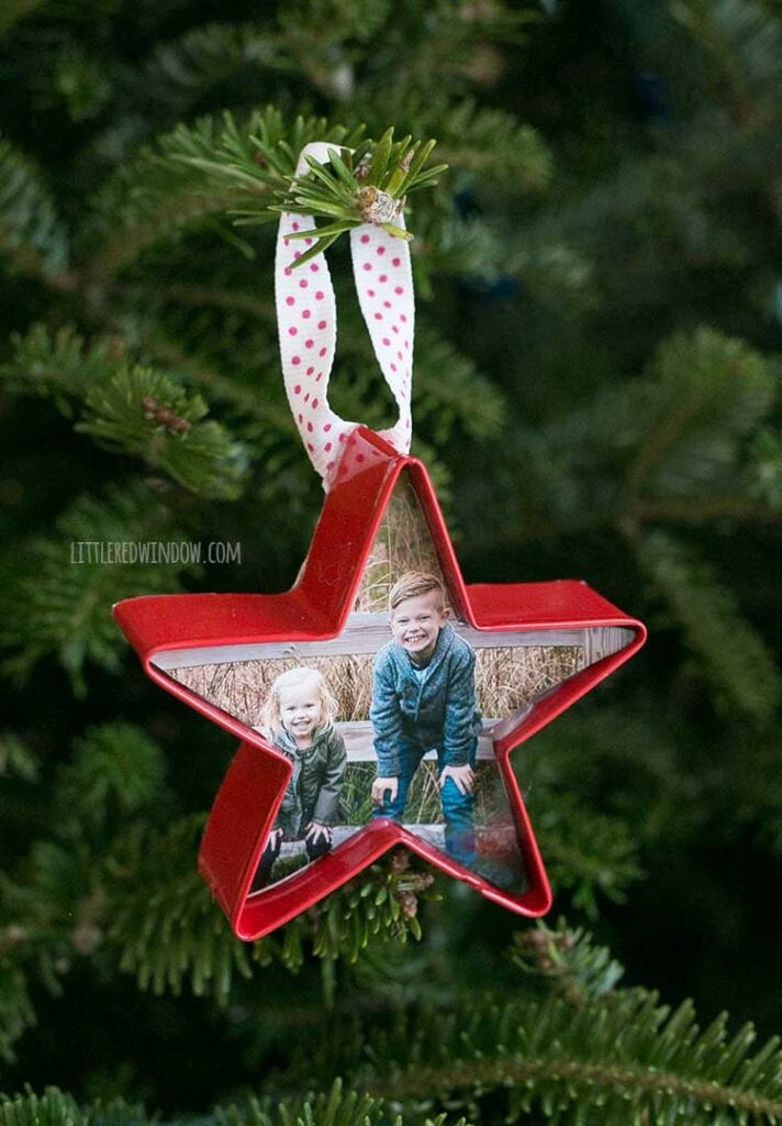 close up of a red cookie cutter ornament with a photo of two children in side and a ribbon hanging loop hanging from the green branch of a Christmas tree