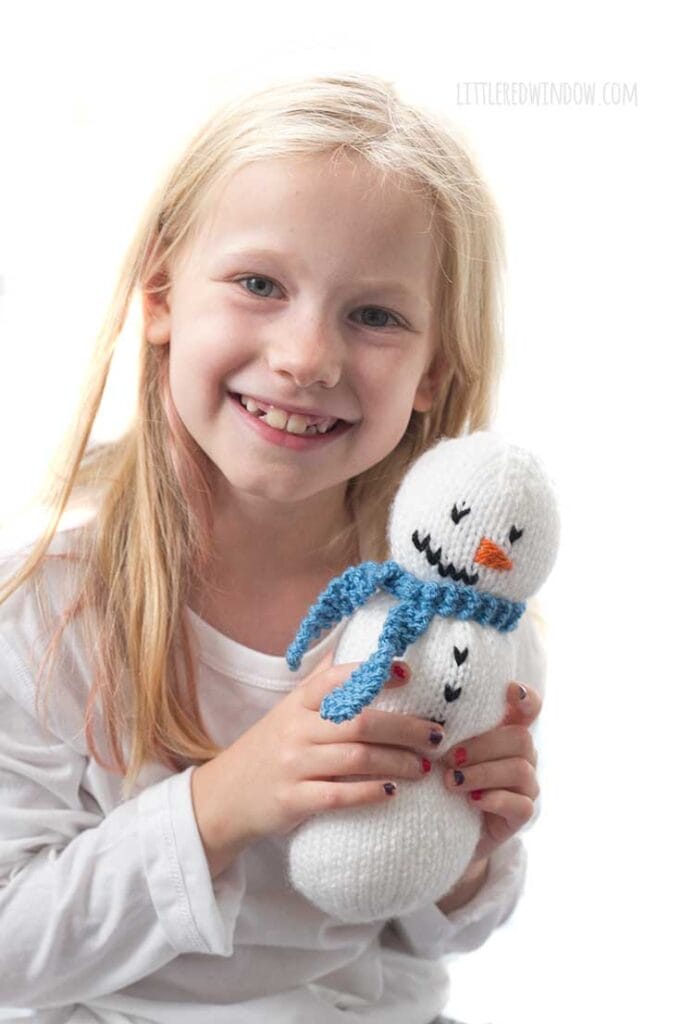 little blond girl smiling and holding a knit snowman with blue knit scarf on it