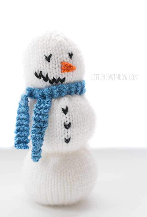 front view of cute white knit showman with black knit button orange yarn nose and a blue knit scarf standing in front of a white background