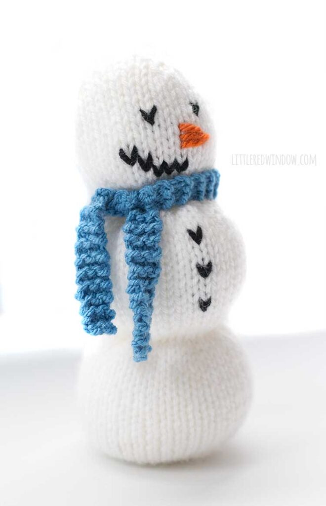 side view of cute white knit showman with black knit button orange yarn nose and a blue knit scarf standing in front of a white background