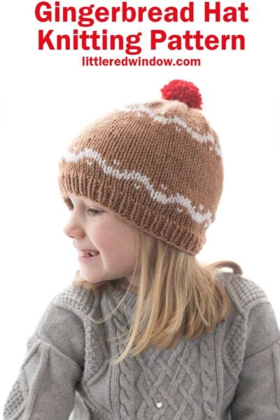 girl in gray sweater wearing a medium brown hat with white designs that look like gingerbread and a red pom pom on top looking off to the left