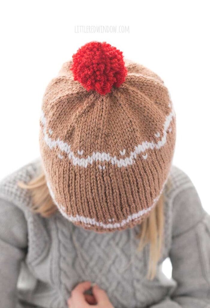 top view of girl wearing a medium brown knit hat with white designs on it that look like gingerbread and a red pom pom on top