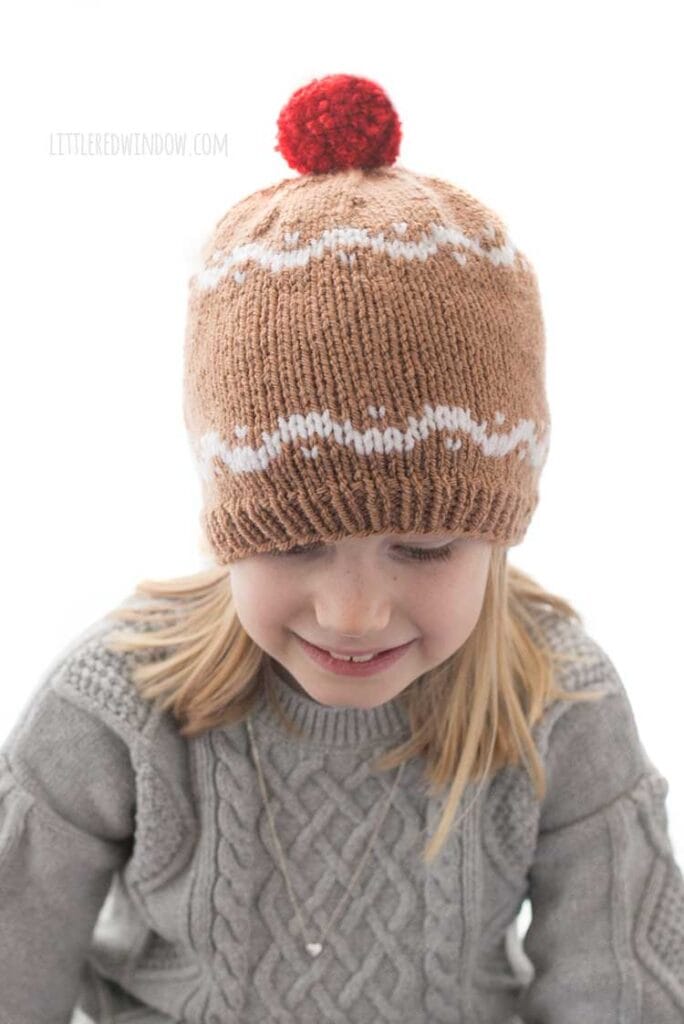 girl in gray sweater wearing a medium brown hat with white designs that look like gingerbread and a red pom pom on top looking down
