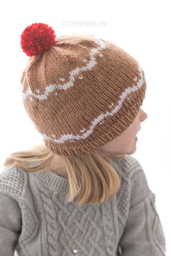 girl in gray sweater wearing a medium brown hat with white designs that look like gingerbread and a red pom pom on top looking off to the right