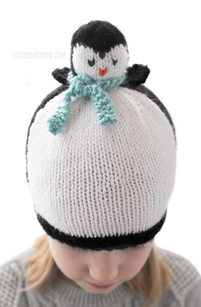 Blonde girl in a black and white hat that looks like a penguin wearing a blue scarf looking down at her lap in front of a white background