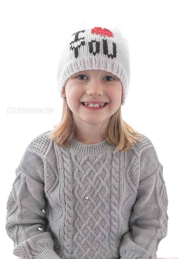 girl in gray sweater dress wearing a white knit hat with the words I heart you where the heart shape is red in front of a white background smiling and looking at the camera