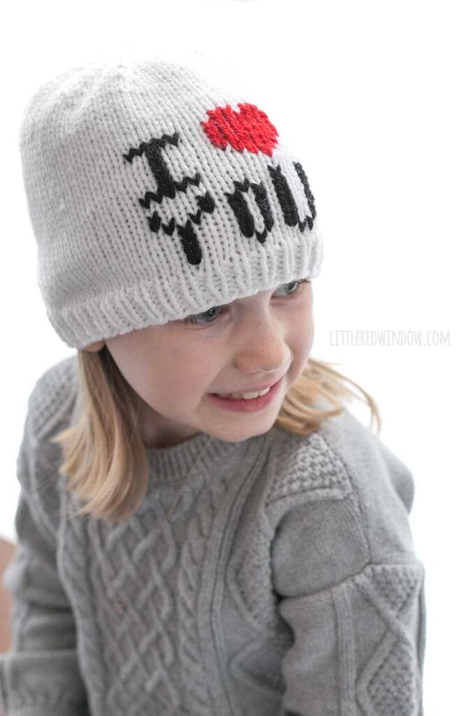 girl in gray sweater dress wearing a white knit hat with the words I heart you where the heart shape is red in front of a white background looking back over her left shoulder