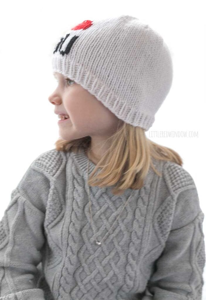 girl in gray sweater dress wearing a white knit hat with the words I heart you where the heart shape is red in front of a white background looking off to the left with a smile