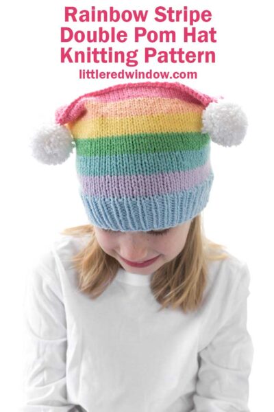 girl in white shirt wearing a rainbow striped hat with two white pom poms on the top corners looking down at her lap