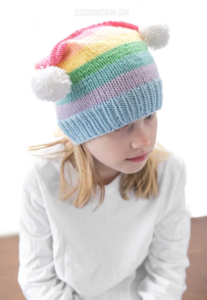 girl in white shirt wearing a rainbow striped hat with two white pom poms on the top corners looking down and to the right