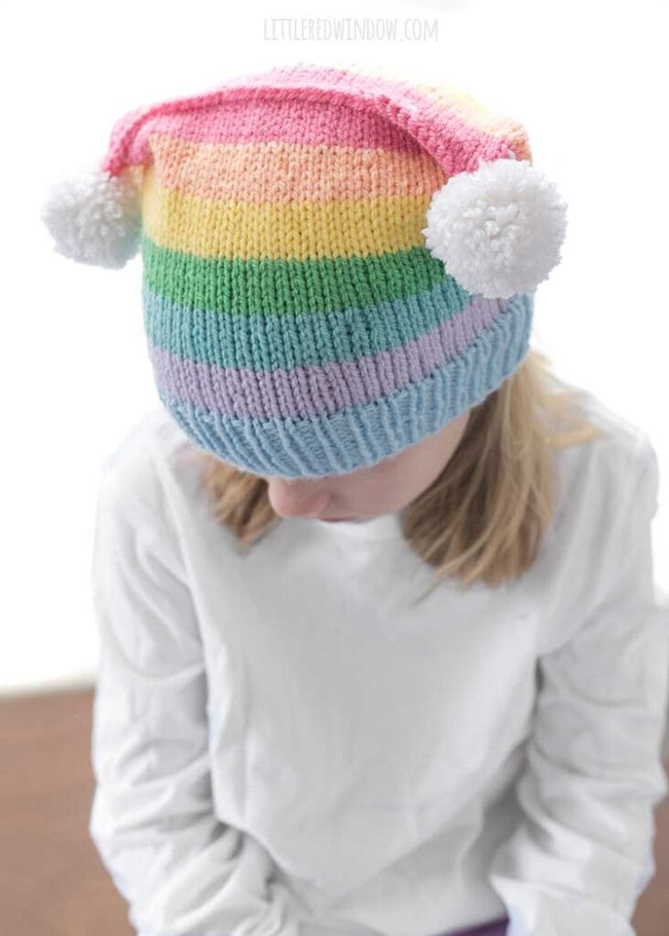 girl in white shirt wearing a rainbow striped hat with two white pom poms on the top corners looking down and to the left