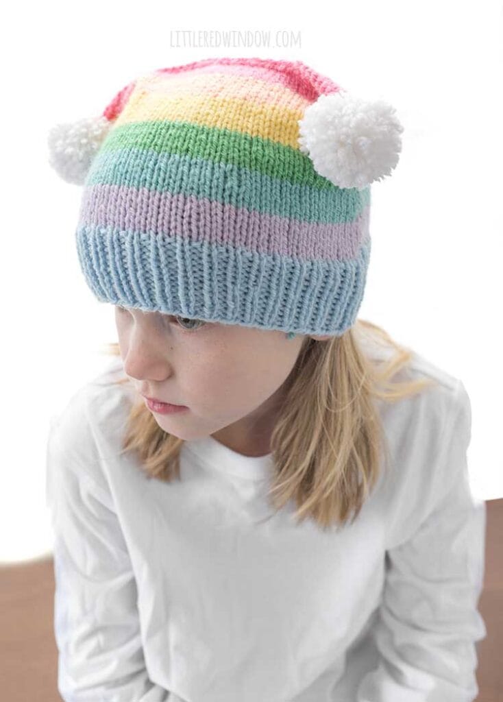 girl in white shirt wearing a rainbow striped hat with two white pom poms on the top corners looking off to the left