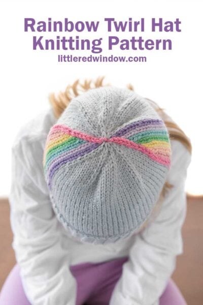 child in white shirt leaning forward towards the camera so you we can see the top of a light blue knit hat with vertical rainbow stripes twirling up towards the top in front of a white background