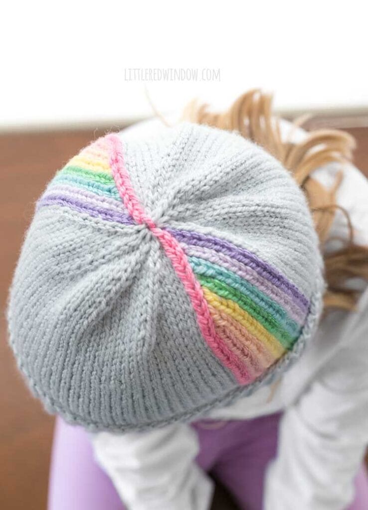 top view of child in white shirt leaning forward towards the camera so you we can see the top of a light blue knit hat with vertical rainbow stripes twirling up towards the top in front of a white background