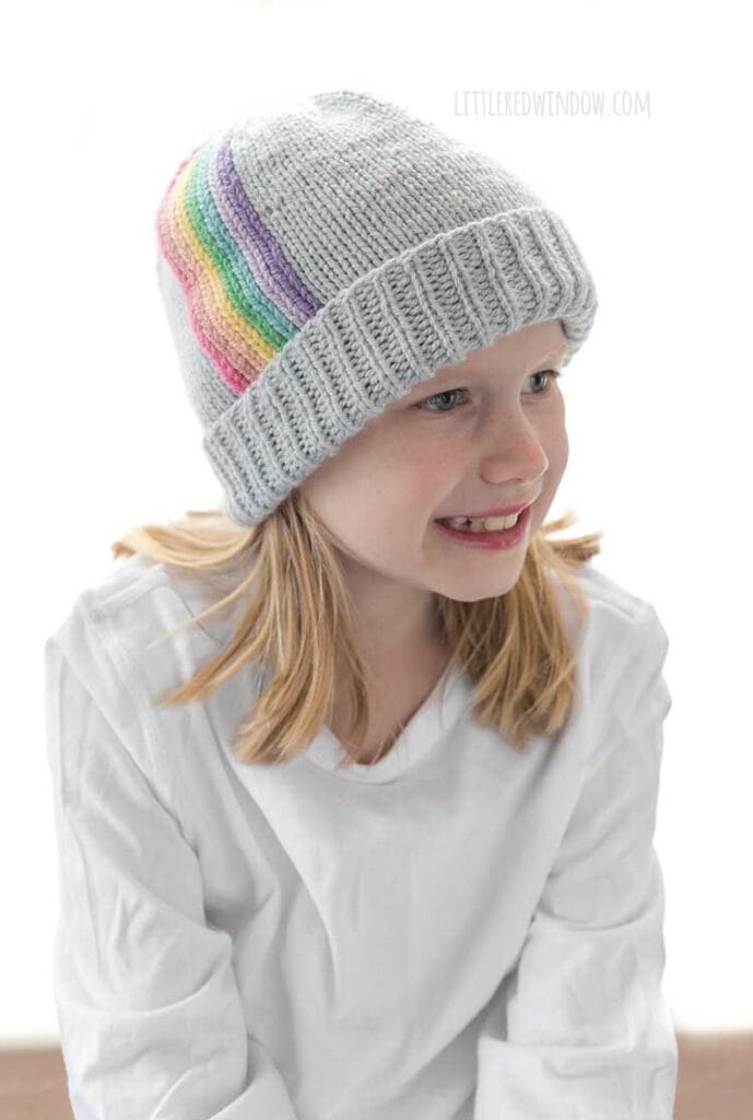 Smiling child in white shirt wearing light blue knit hat with folded brim and vertical rainbow stripes going up towards the top of the hat looking off to the right