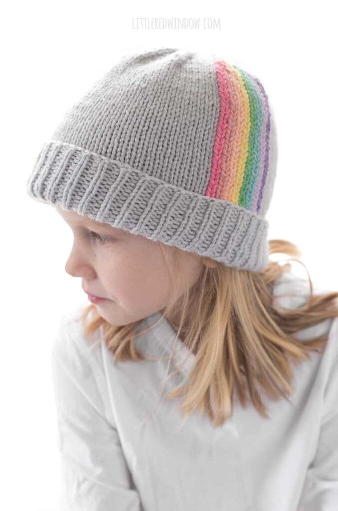 Smiling child in white shirt wearing light blue knit hat with folded brim and vertical rainbow stripes going up towards the top of the hat looking off to the left
