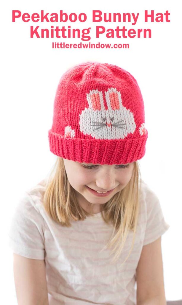 smiling girl in striped short sleeved shirt wearing a hot pink knit hat that has a white bunny face and paws peeking out over the folded brim looking down at her lap