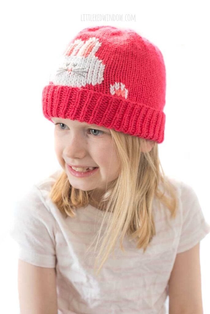 smiling girl in white and light pink striped short sleeved shirt wearing a hot pink knit hat that has a white bunny face and paws peeking out over the folded brim looking over to the left