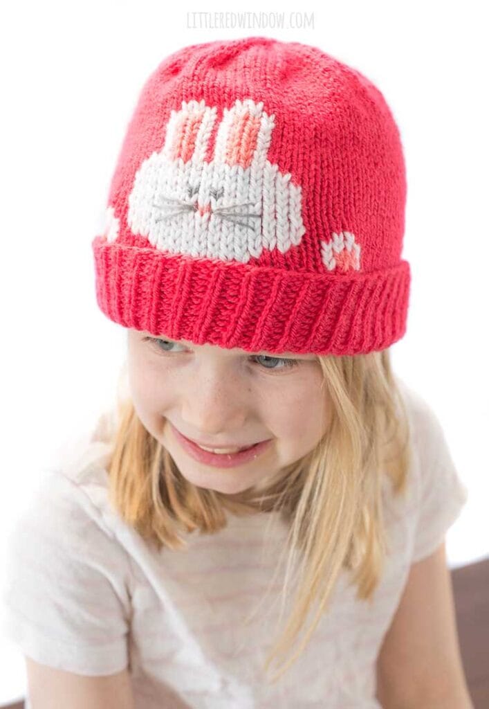 view from slightly above smiling girl in striped short sleeved shirt wearing a hot pink knit hat that has a white bunny face and paws peeking out over the folded brim looking slightly to the left