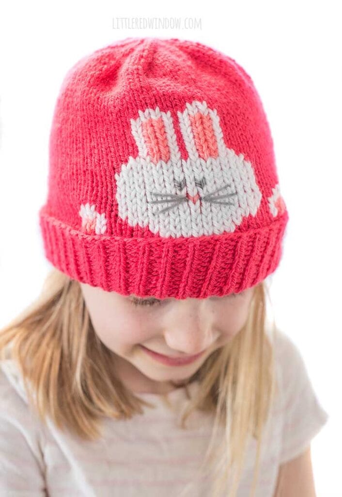 view from straight on of smiling girl in striped short sleeved shirt wearing a hot pink knit hat that has a white bunny face and paws peeking out over the folded brim looking down
