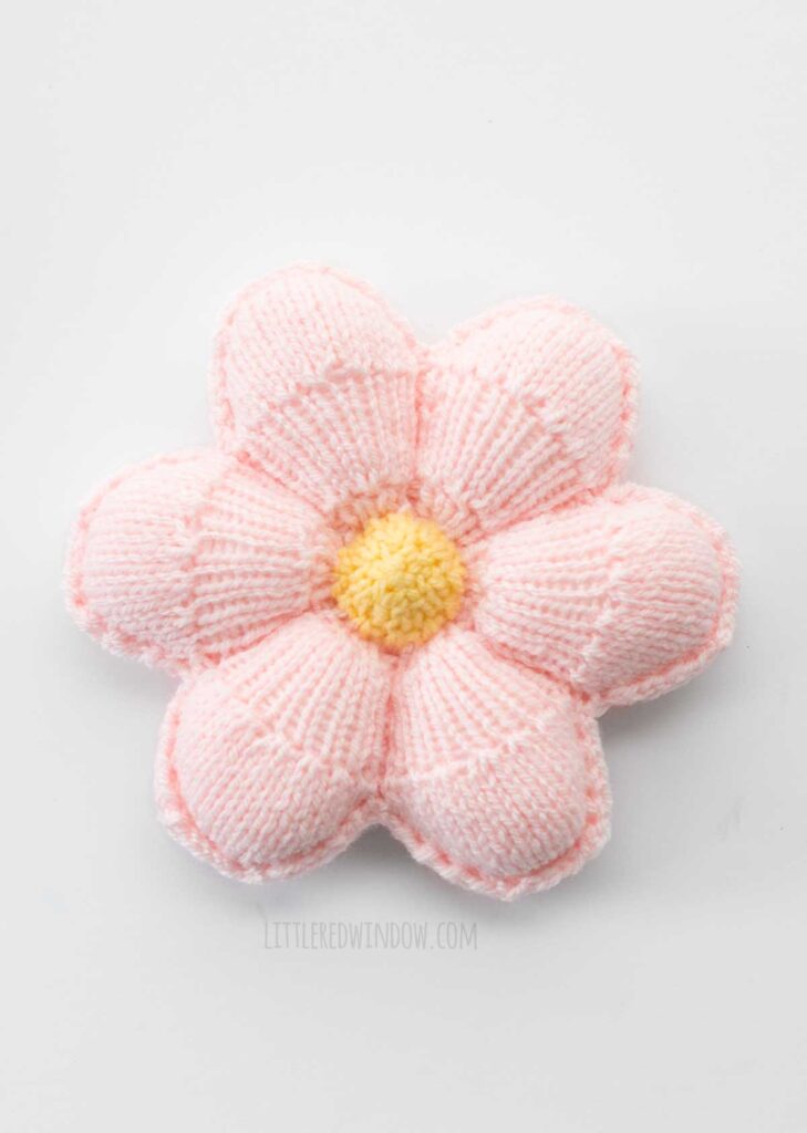 view from above of of small knit flower pillow with light yellow center and light pink petals on a white background
