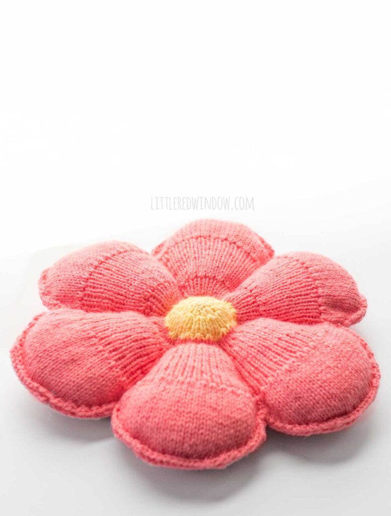 angled view of large knit flower pillow with light yellow center and medium pink petals on a white background