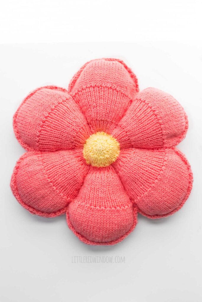 top view of large knit flower pillow with light yellow center and medium pink petals on a white background
