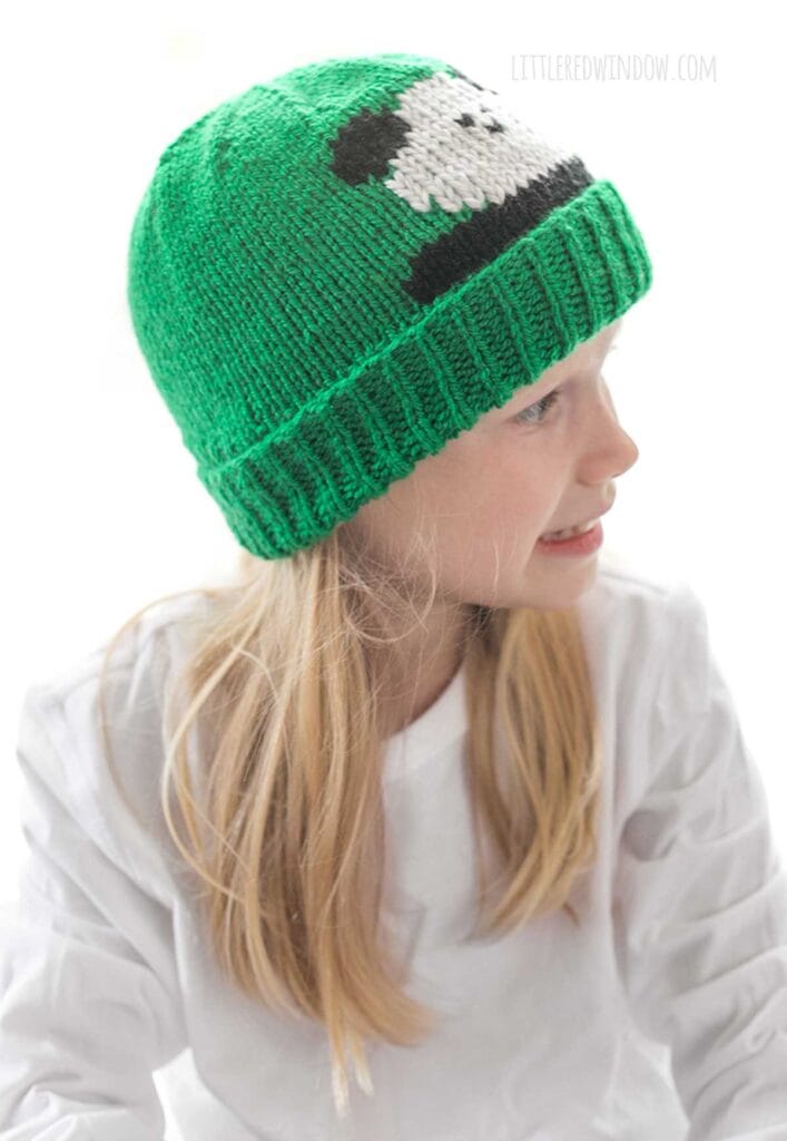 Closeup of Blond girl in white shirt wearing a kelly green knit hat with folded ribbed brim that has a panda face peeking out over the brim while she is smiling and looking down