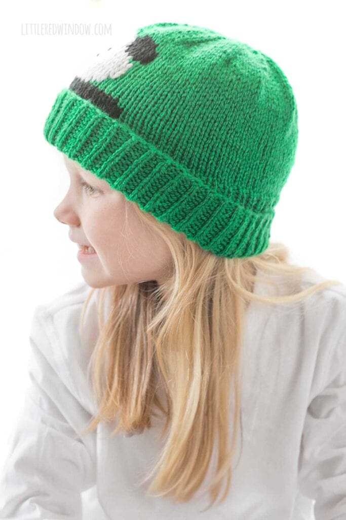 Blond girl in white shirt wearing a kelly green knit hat with folded ribbed brim that has a panda face peeking out over the brim while she is smiling and looking off to the left