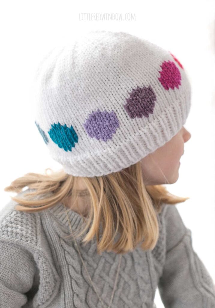 child in gray cable knit sweater wearing a white knit hat with cool rainbow colored large polka dots in a line around the middle looking off to the right