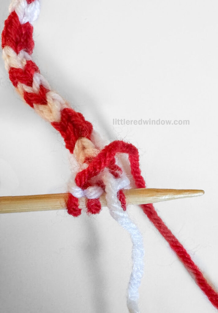 A close-up of a wooden knitting needle holding several stitches of candy cane icord made from red, white, yarn, with a loose white yarn strand trailing to the side on a white background.