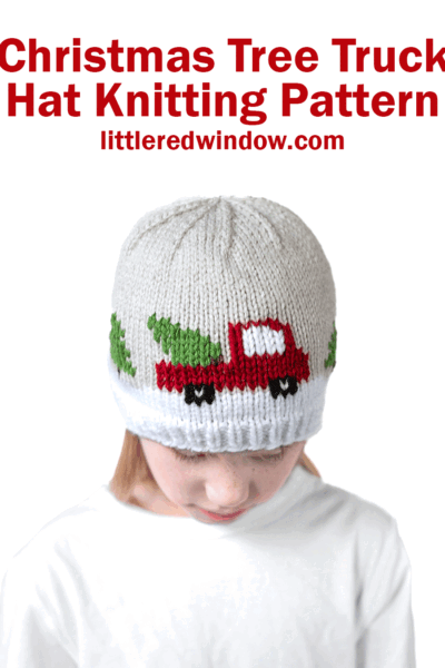 A child wears a cozy knitted hat featuring a christmas tree truck motif—complete with a red truck bringing home a green Christmas tree. Above, the text reads, "Christmas Tree Truck Hat Knitting Pattern, littleredwindow.com.