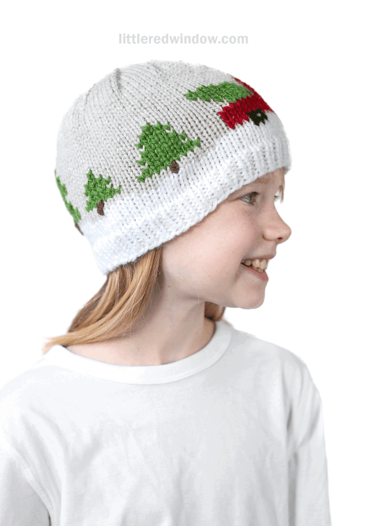 A smiling child wearing a white knit hat with green Christmas trees and a red Christmas tree truck design, facing to the right, in a plain white shirt against a white background.