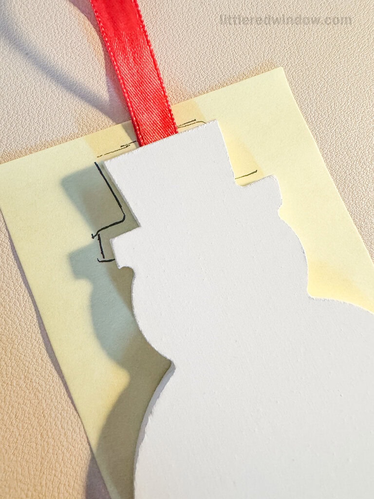 A white snowman-shaped cutout with a red ribbon is placed on a yellow sticky note, casting a shadow. Simple black lines are drawn on the sticky note, to trace the top hat.