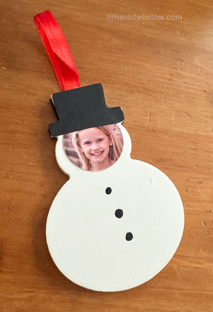 A handmade snowman ornament with a black hat, red ribbon, and a photo of a smiling child’s face as the snowman’s head, lying on a wooden surface.