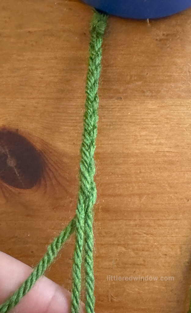 A close-up of a green yarn braid on a wooden surface, with three strands at the end being separated by a hand.
