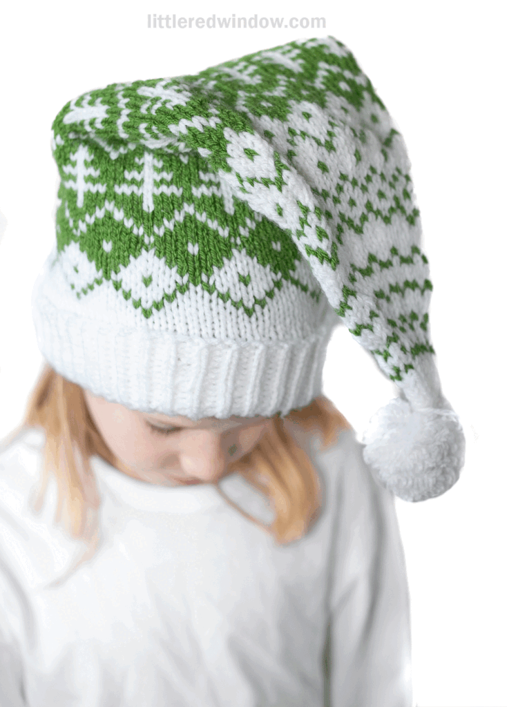 A child wearing an evergreen stocking cap with a white and green winter pattern of trees and geometric designs. The hat, topped with a pom-pom, rests over blonde hair as the child in a white shirt looks down.