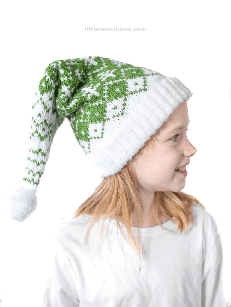 A young child in a white shirt smiles while wearing an evergreen stocking cap with a diamond pattern and a white pom-pom. The background is plain white.