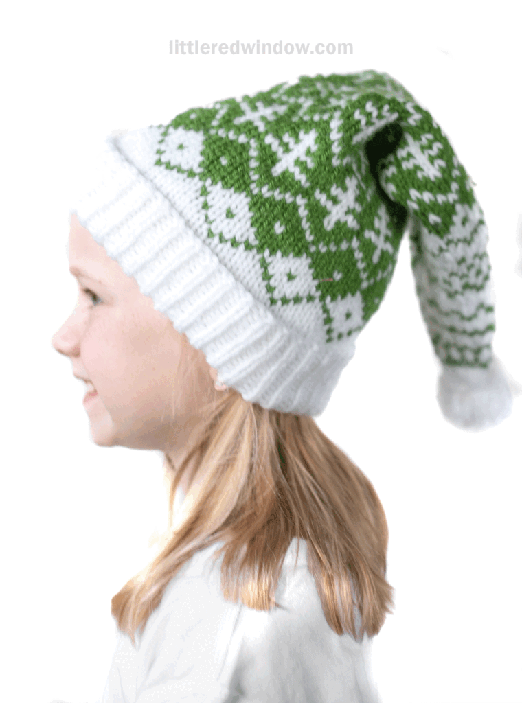 A young girl in profile wears an evergreen stocking cap with a fair isle pattern and a white pom-pom, smiling against a white background.